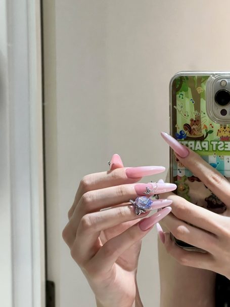 A hand with long pink nails holding a ring, reflecting a smartphone in the background.