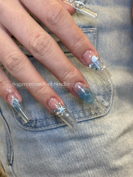 Close-up of a hand with long, clear acrylic nails featuring blue gradients and glitter accents.